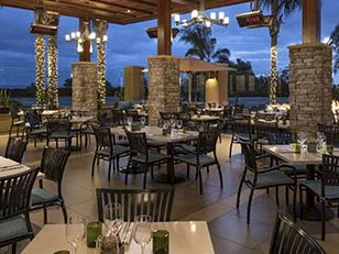An outdoor dining patio with many empty tables, leather chairs, soft lights, stone pillars, and palm trees under a twilight sky, elegant and tranquil.