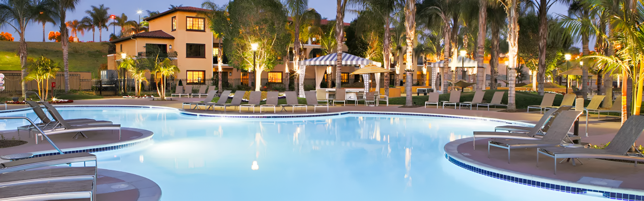 A resort-style pool area at dusk with palm trees, lounge chairs, and a few buildings in the background, lights glowing warmly.