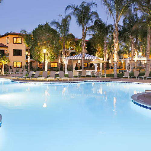 A resort-style pool area at dusk with palm trees, lounge chairs, and a few buildings in the background, lights glowing warmly.